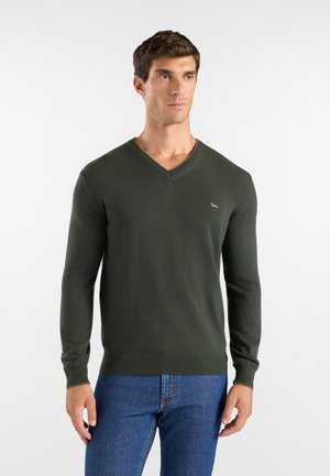 Uomo che indossa un maglione verde scuro scollo a V e jeans blu, in piedi contro uno sfondo chiaro uniforme.