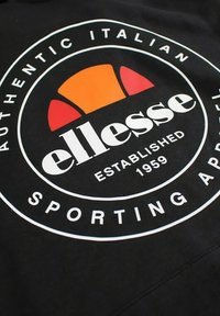 Un tissu noir présente un logo circulaire avec le mot « ellesse » en texte blanc gras, des accents orange et rouge rappelant un soleil, et la mention « AUTHENTIC ITALIAN » au-dessus.