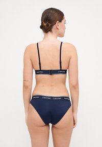 Navy-blauer Bikini aus glattem Material mit verstellbaren Trägern und einem Markenbund mit goldenen Akzenten. Rückansicht gezeigt.