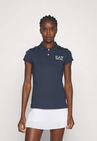 Cămașă polo de culoare navy cu mâneci scurte, cu guler cu nasturi și logo alb brodat. Asortată cu o fustă albă plisată.