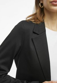 Černá blazer s strukturovaným designem, texturovanou látkou a zapínáním na jedno tlačítko. Obsahuje klopu s zářezem a přiléhavé rukávy.