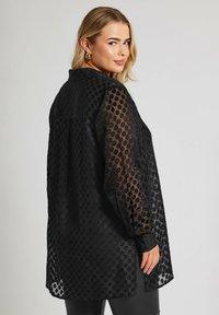 Czarna przezroczysta bluzka w kropki, z długimi rękawami, mankietami na guziki i kołnierzykiem. Prosty krój z rozcięciami po bokach, wykonana z lekkiego materiału.