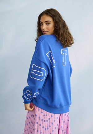 Jonge vrouw met krullend haar, gekleed in een oversized blauwe trui met witte letters en een roze rok met patroon, kijkt over haar schouder.