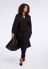 Abrigo largo negro con interior de satén, combinado con una blusa negra con un cuello decorativo y pantalones negros ajustados. Tacones negros puntiagudos.