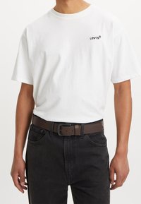 Biała bawełniana koszulka z okrągłym dekoltem i czarnym logo "Levi's", zestawiona z ciemnymi jeansami i brązowym skórzanym paskiem z metalową klamrą.