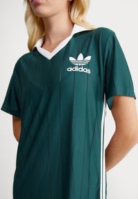 Zaļa sporta krekls ar baltu apkakli, ar vertikālām melnām svītrām un Adidas logo kreisajā krūtī. Īsas piedurknes.
