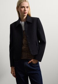 Veste en mélange de laine noire avec un design à col, des boutons sur le devant et des poches latérales, portée sur un haut en maille marron et un jean en denim foncé.
