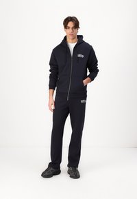 Marineblaue Zip-Hoodie mit frontalen Taschen, kombiniert mit passenden Hosen. Aus weichem Stoff mit dezentem Logodetail. Schwarze Sneakers.
