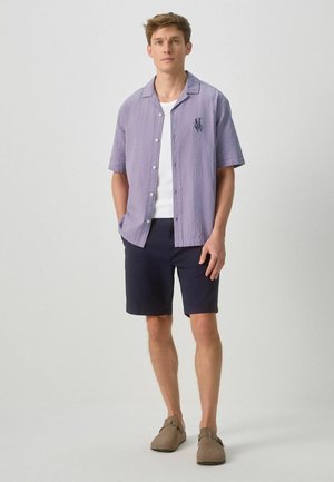 Mężczyzna stoi, ubrany w jasnofioletową koszulę na krótki rękaw, zapinaną na guziki, na białym t-shircie, granatowe szorty i brązowe buty wsuwane.