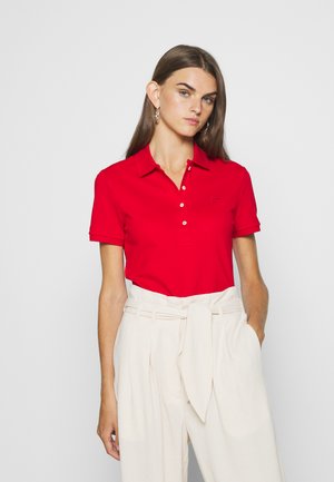 Polo rouge avec col et manches courtes, comportant une patte de boutonnage à trois boutons et un petit logo sur la poitrine, associé à un pantalon beige clair.