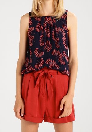 Kobieta w granatowej bluzce bez rękawów z czerwonym wzorem liści oraz czerwonych szortach z wysokim stanem i wiązaną kokardą w pasie.