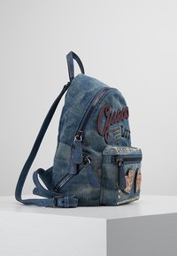 Mochila de mezclilla con texto bordado "Guess", un bolsillo frontal con cremallera adornado con corazones y correas ajustables con acabado texturizado.