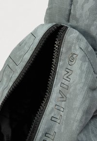 Polstret grå jakke med teksturert stoff, med en svart glidelås og trykt tekst på forsiden. Innsiden viser en mørk foret.