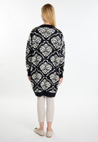 Přes veliký pletený cardigan v černé a béžové barvě s intricate květinovým vzorem, pletené manžety a lem. Nosený přes světlé béžové kalhoty.