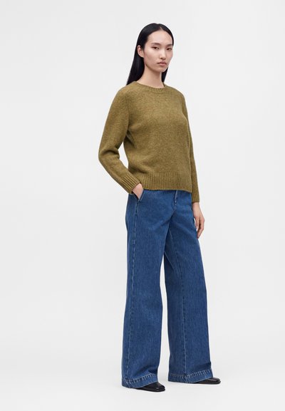Femme aux longs cheveux noirs portant un pull vert olive, un jean bleu à jambes larges et des chaussures noires, debout avec une main dans la poche.