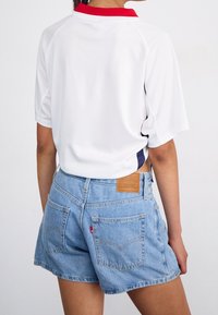 Chemise blanche à manches courtes avec un col rouge, associée à un short en denim bleu clair avec des poches arrière et une étiquette en cuir beige.