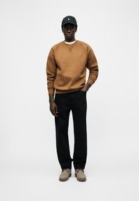 Άνδρα ς που φοράει καφέ φούτερ, μαύρο παντελόνι, μπεζ παπούτσια και μαύρο καπέλο, στέκεται μπροστά σε απλό λευκό φόντο.