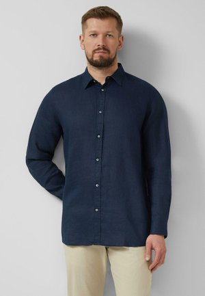 Mann mit Bart, der ein dunkelblaues langärmeliges Hemd und beige Hose trägt, steht vor einer hellgrauen Wand.