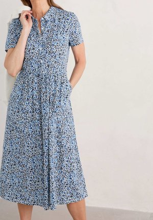Femme portant une robe midi boutonnée à manches courtes à fleurs bleues et blanches avec des poches, tenant une veste blanche sur son épaule.