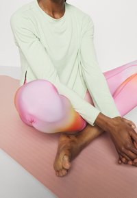 Persona seduta con le gambe incrociate su un tappetino da yoga rosa, indossa una maglietta a maniche lunghe verde chiaro e leggings rosa sfumati, con le mani unite sopra il piede.