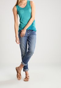 Tyrkysový top bez rukávů s květinovými vzory, doplněný světle modrými ohrnutými džíny a hnědými sandály na pásky. Jednoduchý a ležérní design.