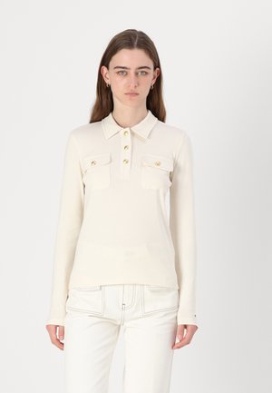 Polo a maniche lunghe color crema con colletto, chiusura con bottoni dorati e due tasche a pattina. Tessuto liscio, vestibilità sagomata, abbinato a pantaloni bianchi.