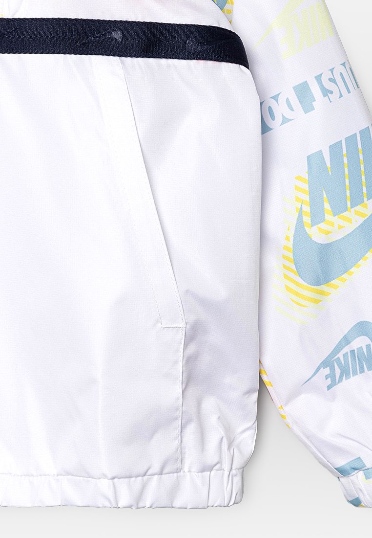 Veste blanche avec une bande d'accent marine. Présente une marque Nike en bleu clair et jaune, avec un tissu léger et texturé et des poignets élastiques.