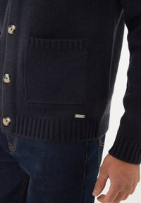Navy gebreid cardigan met een geribde zoom en manchetten, voorzien van een grote zak en knopen van crème-kleurig materiaal. Zilveren logo-accent.