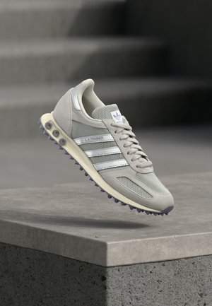 Hellgrauer Adidas L.A. Trainer Sneaker mit weißen Streifen, der über einer Betonabsatzkante schwebt vor einem verschwommenen Treppenhintergrund.