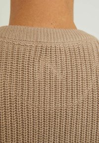 Beige knitted sweater with a ribbed texture, featuring a subtle embroidered logo detail on the back neckline. Simple and minimal design.