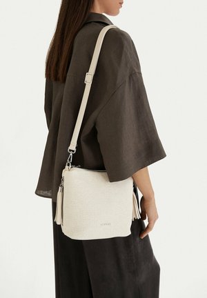 Donna che indossa una camicia larga scura e una gonna, con una borsa a tracolla color crema con trama, nappine e dettagli in argento sulla spalla.