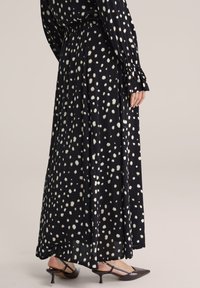 Melna maksi kleita ar baltām neregulārām polka punktu apdrukām, elastīgu vidukli, garām piedurknēm ar volāna aprocēm, kopā ar melniem slingbeka apaviem ar zemu papēdi.