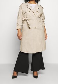 Beige gestreepte trenchcoat met dubbele knopen, riem, schouderepauletten en een relaxte pasvorm, gecombineerd met zwarte wijde broek en hakken.