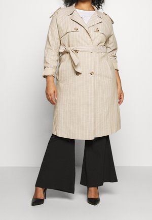 Trench coat a rayas beige con botones de doble botonadura, cinturón, hombreras y un corte relajado, combinado con pantalones de pierna ancha negros y tacones.