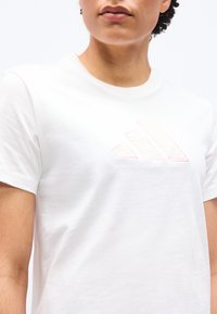 Κοντινή λήψη ενός ατόμου που φοράει λευκό κοντομάνικο μπλουζάκι με στρογγυλή λαιμόκοψη και διακριτικό ανάγλυφο τριγωνικό λογότυπο στο στήθος.