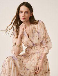 Joven con cabello largo y castaño que lleva un vestido beige claro con estampado floral, mangas abullonadas y un cinturón a juego, posando con la mano cerca del rostro.