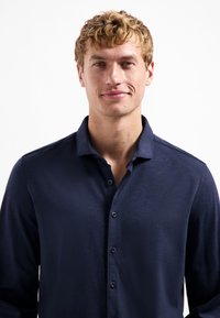 Blonde man met kort krullend haar, gekleed in een marineblauwe button-up shirt, die naar voren kijkt met een lichte glimlach tegen een witte achtergrond.