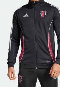 Chaqueta deportiva negra con cremallera, acentos rojos en los laterales y rayas blancas. Presenta la etiqueta y el logo "AEROREADY" en el pecho. El material es suave.