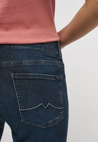 Jeans en denim bleu foncé avec une finition texturée. Présente un design de poche arrière avec une courbe distincte et une étiquette en cuir noir de marque.