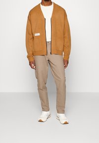 Brun stickad kofta med dragkedja framtill, två fickor och ribbade muddar, bärs över en vit skjorta och kombineras med beige chinos och vita sneakers.