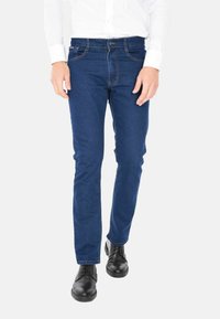 Uomo che indossa jeans slim-fit blu, camicia bianca e scarpe eleganti nere, in piedi contro uno sfondo bianco.