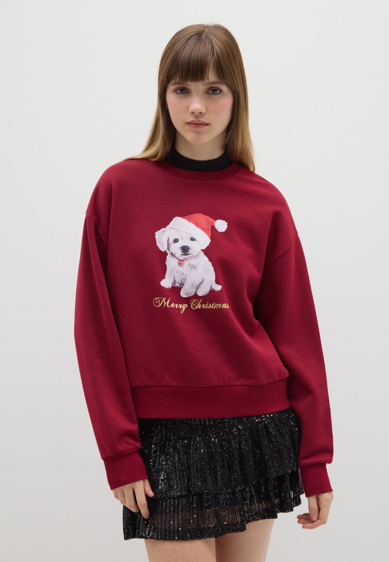 Rode sweatshirt met een witte puppy in een kerstmanhoed, met "Zalig Kerstfeest" in goud eronder gedrukt. Draag je over een zwarte paillettenrok.