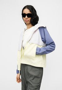 Γυναίκα που φοράει μαύρα γυαλιά ηλίου, αδιάβροχο με έντονα χρώματα με μωβ μανίκια, μπεζ κορμό και γκρι παντελόνι, στέκεται μπροστά σε λευκό φόντο.