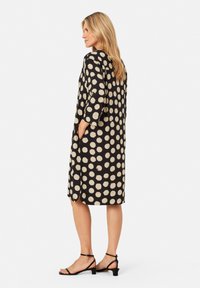 Schwarzes Kleid mit großen beigen Punkten, drei Viertel-Ärmeln, seitlichen Taschen und geradem Schnitt mit seitlichen Schlitzen, kombiniert mit schwarzen Sandalen.