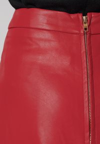 Jupe en cuir rouge avec une fermeture éclair à l'avant et une texture lisse. Présente une taille ajustée et des coutures décoratives, mettant en valeur le matériau brillant.