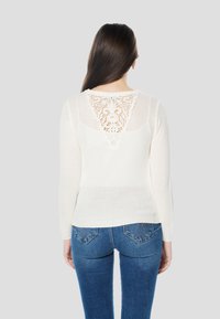 Witte gebreide trui met een kanten patroon in V-vorm op de achterkant, met lange mouwen en een ontspannen pasvorm, gecombineerd met een blauwe spijkerbroek.