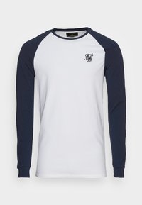 Valkoinen pitkähiha raglan-paita, jossa on tummansiniset hihat ja kaulus, ja vasemman rinnan kohdalla pieni musta brodeerattu monogrammi.