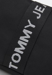 Black fabric pouch featuring white embroidered text "TOMMY JEANS," with a smooth texture and a rectangular shape. Zipper closure visible.