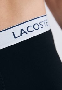 Czarne bokserki posiadają białą elastyczną talię z napisem "LACOSTE" w granatowym kolorze. Gładka tkanina, dopasowany krój z detalami szwów.