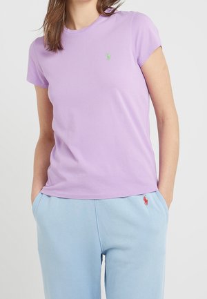 Kobieta ubrana w jasnofioletowy krótki rękaw t-shirt oraz jasnoniebieskie spodnie, z rękami w kieszeniach, pokazująca haftowane logotypy na obu ubraniach.